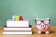 The letters FSA sitting on top of a stack of books, next to a piggy bank wearing glasses [Image by creator Tierney from AdobeStock]